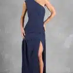 A woman stands against a gray textured backdrop wearing a dark blue, one-shoulder evening gown made with a sewing pattern, featuring a thigh-high slit on the left side. She has her hair styled up and accessorizes with dark high-heeled shoes. Her left hand rests on her hip while the right hand hangs by her side.