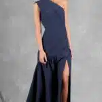 A woman stands in a full-length, asymmetrical, navy blue gown made with a sewing pattern, featuring one shoulder strap and a high slit on the left side, revealing her left leg. She wears metallic silver heels and has her hair styled in a low updo. The background is a textured gray.