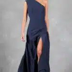 A person is standing against a gray textured background. They are wearing a navy blue patron de couture robe with a high slit on one side, revealing part of their leg. The individual has light skin and their hair is tied back. They are wearing silver open-toe sandals.