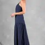 A woman stands against a gray textured background, wearing a navy blue, one-shoulder, floor-length dress made with a sewing pattern. She has her hair tied back in a low bun and is wearing clear, high-heeled shoes. The dress drapes elegantly, showcasing a slightly gathered waist as she poses with her body facing left.
