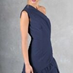A woman with brown hair in an updo wears a navy blue, one-shoulder gown made with a sewing pattern. She stands against a gray backdrop, looking down with her right hand touching the side of her dress. The gown features a fitted bodice and a flowing skirt with a slight train.