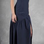 A woman wearing an elegant, sleeveless, dark navy blue gown made with a sewing pattern poses against a gray, textured backdrop. The floor-length gown features a high slit on the side, revealing part of her leg. She stands with her left arm relaxed by her side, highlighting the gown's fitted bodice and flowing lower skirt.