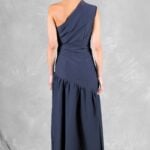 A woman with her hair in a bun is seen from the back, wearing a navy blue, one-shoulder dress with an asymmetrical neckline. The long dress, made with a sewing pattern, features a gathered detail at the waist and a slight flare. The background is a textured, light gray surface with fabric on the floor.