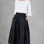 Une femme se tient avec confiance sur un fond gris. Elle porte un chemisier blanc ample à manches trois-quarts et une jupe midi noire taille haute créée avec patron de couture jupe femme. Elle a nonchalamment les mains dans les poches et porte des sandales à talons hauts beiges à lanières. Ses cheveux sont tirés en arrière.
