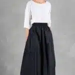 Une femme aux cheveux noirs attachés en arrière se tient sur un fond gris. Elle porte un chemisier blanc ample à manches trois-quarts et une longue jupe trapèze noire, créée avec patron de couture jupe femme, qui lui arrive aux chevilles. Ses mains sont dans les poches de sa jupe alors qu'elle regarde à sa gauche. Elle porte également des sandales blanches.
