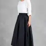 Une femme se tient sur un fond gris, vêtue d'un chemisier blanc ample à manches trois-quarts et d'une jupe trapèze noire taille haute créée avec une jupe femme patron de couture qui tombe sous le genou. Elle a les cheveux châtain clair tirés en arrière et porte des sandales à talons de couleur claire. Elle regarde la caméra avec confiance.