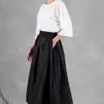 Une femme aux cheveux attachés se tient de côté sur un fond gris. Elle porte un chemisier blanc à manches jusqu'aux coudes et une jupe noire, créée avec patron de couture, qui arrive à mi-mollet. Ses sandales blanches à talons hauts complètent sa tenue. Sa main gauche est dans sa poche.