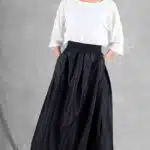 Une jeune femme se tient sur un fond gris et regarde à sa droite. Elle porte un chemisier blanc ample et une jupe noire fluide taille haute créée avec une jupe femme patron de couture, les mains dans les poches de la jupe. Ses cheveux sont attachés en chignon bas. Elle complète la tenue avec des talons ouverts et le look général est élégant et minimaliste.