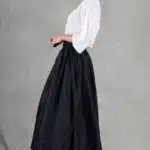 Une femme se tient sur un fond gris uni, vêtue d'un chemisier blanc à manches longues et d'une jupe midi trapèze noire taille haute créée avec patron de couture. Elle porte des talons à lanières blancs et a les cheveux coiffés dans un chignon soigné. Elle est dans une pose de profil, regardant par-dessus son épaule vers la caméra.