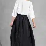 Une femme se tient face à face, exhibant le dos de sa tenue. Ses cheveux sont soigneusement attachés en chignon. Elle porte un chemisier blanc ample à manches longues et une jupe noire taille haute mi-longue créée avec patron de couture. Des sandales blanches à talons ouverts complètent son look sur le fond gris uni.