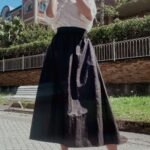 Une femme se tient dehors devant un bâtiment en brique, vêtue d'un chemisier blanc rentré dans une longue jupe noire confectionnée à l'aide d'un patron de couture jupe femme et de talons hauts beiges. Elle a une main sur le cou et regarde légèrement vers le haut. L'arrière-plan présente un sol pavé hexagonal, un banc blanc et de la verdure autour du bâtiment.