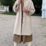 BRISE long coat sewing pattern in a full-length outdoor photo: cream textured coat with front buttons worn over brown clothing and tall ivory boots.
