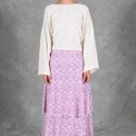 Back view of the KELIS batwing-sleeve top sewing pattern, showing its relaxed cream bodice, long sleeves and gently gathered waist above a pink floral skirt.