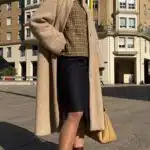 A woman stands outside in front of a large, multi-story building. She is wearing a long beige coat made with a sewing pattern over a plaid top, a dark knee-length skirt, and burgundy ankle boots. She carries a tan handbag and wears sunglasses. Her hair is tied back, and she stands with her hands in her coat pockets.