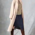 VIVIANE women’s coat sewing pattern: long beige overcoat worn over a checked shirt with a denim skirt and tall brown boots.