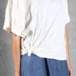 Close-up of the CALYS one-piece top sewing pattern in white, focusing on the softly folded neckline, loose sleeves and knotted fabric near the left hip.