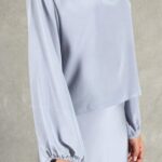 Una persona se encuentra sobre un fondo gris texturizado, vestida con una blusa azul claro de manga larga con cuello en V y una falda a juego. La blusa, hecha con un patrón de costura, tiene mangas relajadas recogidas en las muñecas. La falda recta completa el conjunto. El rostro de la persona no se ve en la imagen.