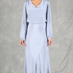 Una mujer, de pie sobre un fondo gris, lleva un conjunto de seda azul claro con mangas largas y escote en V. El conjunto, hecho con un patrón de costura, se extiende por debajo de las rodillas y tiene un ligero brillo. También lleva tacones blancos abiertos. Su cabello oscuro está recogido y tiene una expresión neutral.