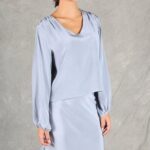 Una mujer de cabello oscuro lleva una blusa sedosa de manga larga de color gris claro combinada con una falda larga a juego, hecha con un patrón de costura. Ella está de pie contra un fondo gris liso, mirando hacia un lado con una expresión neutral. El conjunto presenta sutiles detalles fruncidos en los hombros y las muñecas.
