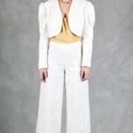 Une femme se tient sur un fond texturé gris. Elle porte un tailleur-pantalon blanc créé avec patron de couture pantalon femme, composé d'une veste courte et d'un pantalon large, associé à un haut jaune moutarde. Ses cheveux sont attachés en arrière et elle les accessoirise avec un collier en or et des talons blancs à bout ouvert.