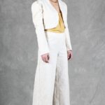 Une personne aux longs cheveux noirs attachés en arrière se tient sur un fond gris. Elle porte un tailleur-pantalon texturé de couleur crème, créé avec patron de couture pantalon femme, avec une veste courte sur un haut jaune moutarde. Elle porte des talons hauts de couleur claire et un collier en chaîne. Ses mains sont détendues à leurs côtés et elle regarde droit devant elle.