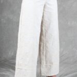 Une personne se tient debout sur un fond gris, représentée du milieu du torse vers le bas. Elle porte un pantalon large texturé de couleur crème avec un motif subtil, probablement créé avec patron de couture pantalon femme, et des talons à lanières beiges. Leurs mains sont placées près de leur taille, une main tenant l’autre poignet.