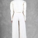 Une personne se tient face à la caméra sur un fond gris. Elle a les cheveux courts et porte un haut blanc à manches longues aux épaules structurées, associé à un pantalon large taille haute créé avec patron de couture pantalon femme. Ils portent également des talons hauts blancs et un collier en or.