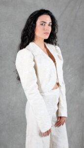 A woman with long, wavy dark hair poses against a gray backdrop. She is wearing an elegant white, bolero jacket made with a bolero jacket pattern and matching high-waisted pants. She gazes confidently at the camera with her hands lightly clasped in front of her.