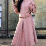 A woman with long, curly black hair stands confidently outside a stone building with red brick accents. She's wearing a matching pink patterned outfit made with a skirt box pleat pattern, consisting of a long-sleeve top and a midi skirt. She pairs the look with cream knee-high boots and sunglasses.