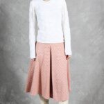 A woman with dark hair stands against a gray backdrop, wearing a white textured long-sleeve top, a knee-length peach and white skirt made with a box pleat pattern, and white knee-high boots. She accessorizes with a gold necklace and has a neutral expression, hands at her sides.