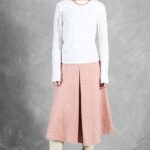 A person with dark hair stands against a gray textured backdrop. They wear a white long-sleeve top with a textured pattern, a pink midi skirt made with a skirt box pleat pattern and small geometric prints, and cream-colored knee-high boots. They accessorize with a gold necklace and wear a neutral expression.