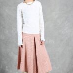 A woman stands in front of a gray backdrop. She wears a white textured long-sleeve top, a pink mid-length pleated skirt made with a box pleat pattern, and knee-high white boots with block heels. Her dark hair is styled short, and she looks to her left with a neutral expression, her arms relaxed by her sides.