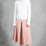 A woman stands against a gray backdrop, wearing a textured white long-sleeve top and a mid-length peach-colored skirt made with a box pleat pattern. She complements the look with white knee-high boots. Her dark hair is styled simply, and she accessorizes with a gold necklace. Her expression is neutral as she gazes off to the side.