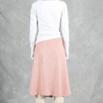 A person with short brown hair stands with their back to the camera against a gray backdrop. They are wearing a white long-sleeve top, a knee-length pink and white skirt made with a box pleat pattern, and tall beige boots. The setting appears to be indoors with studio lighting.
