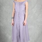 A woman stands against a textured gray backdrop, wearing a lilac, button-front, sleeveless dress that reaches her calves, crafted with a summer dress pattern.