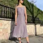 A woman in a lavender midi dress made with a summer dress pattern, stands on a stone-paved sidewalk beside a stone wall and wrought iron fence.