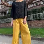 A person wearing white sunglasses, a black long-sleeved top made with a women top sewing pattern, and yellow wide-leg pants poses outdoors on a paved path. They are also wearing black platform sandals. Behind them are residential buildings, a fence, and a tree with bare branches. The day appears sunny.