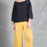 A woman stands against a textured gray backdrop. She wears a black, long-sleeved top crafted with a women top sewing pattern.