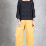 A woman stands against a textured gray backdrop. She wears a black, long-sleeved top crafted with a women top sewing pattern.