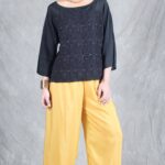 A woman stands against a textured gray backdrop. She wears a black, long-sleeved top crafted with a women top sewing pattern.