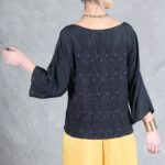 A person with dark hair styled in a bun is standing against a grey background. They are wearing a black long-sleeve blouse made with a women's top sewing pattern and yellow wide-leg pants. Their right arm is slightly raised, and their face is turned away from the camera. They are accessorized with gold bangles and a gold necklace.