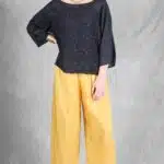 Una mujer se encuentra frente a un fondo texturizado gris, vestida con una blusa negra de manga 3/4 con un patrón texturizado y un pantalón pirata amarillo holgado hecho con patrón de costura. Complementa con un collar dorado, una pulsera dorada y sandalias de cuña negras. Posa con la mano derecha en la cadera, mirando ligeramente hacia la izquierda.