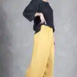 Una mujer se alza sobre un fondo gris y posa con una mano en la cadera. Lleva una blusa negra holgada con mangas anchas, combinada con pantalones palazzo de cintura alta de color amarillo mostaza hechos con un patrón de costura personalizado. También lleva sandalias de plataforma negras y una pulsera dorada, con el cabello oscuro recogido en un moño.