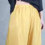 Una persona está vestida con una blusa negra texturizada y pantalones amarillos holgados de cintura alta, hecho con patrón de costura. Tiene un brazalete de oro en la muñeca izquierda y su mano izquierda se coloca sobre la cintura del pantalón, levantando ligeramente la blusa. El fondo es un gris neutro.