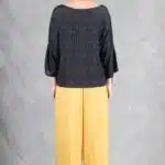 Persona de pie con la cámara mirando hacia atrás contra un fondo gris. Llevan un top negro con diseño texturizado, pantalón pirata holgado hecho con patrón de costura en color amarillo y sandalias de tacón negras. Llevan el cabello recogido hacia atrás y tienen algunas joyas que incluyen un collar, una pulsera y un anillo grande en la mano izquierda.