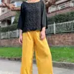 Una mujer se encuentra al aire libre en una acera pavimentada, con gafas de sol, una blusa negra con textura y un pantalón pirata amarillo brillante hecho con patrón de costura. Tiene el pelo corto y también lleva zapatos de plataforma negros. Detrás de ella hay arbustos verdes, un edificio de ladrillo y árboles sin hojas bajo un cielo nublado.