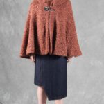 Woman wearing a cozy brown cape, crafted with a ladies cape pattern, featuring a rounded collar and a decorative brooch for closure.