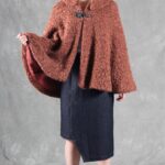 Woman wearing a cozy brown cape, crafted with a ladies cape pattern, featuring a rounded collar and a decorative brooch for closure.