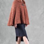 Woman wearing a cozy brown cape, crafted with a ladies cape pattern, featuring a rounded collar and a decorative brooch for closure.