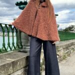 Woman wearing a cozy brown cape, crafted with a ladies cape pattern, featuring a rounded collar and a decorative brooch for closure.